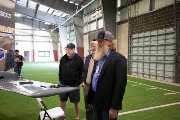 Three men stand indoors on artificial turf, observing a display with aviation-related decals. Two wear caps and casual jackets, while the third wears a blazer and has a long beard. The setting appears to be a large industrial or sports facility.