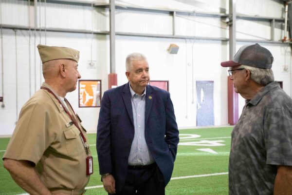 Three men stand and talk on an indoor football field: one in a tan military uniform, one in a suit, and one in a gray shirt and cap. They appear to be having a conversation.