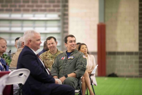 A group of people, including a man in a military flight suit, sit on white chairs indoors, listening and smiling during what appears to be a formal event. Some wear uniforms; others are in civilian attire.