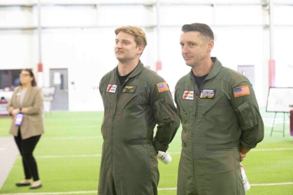 Two men in green flight suits stand side by side indoors on artificial turf. Both have name and American flag patches on their suits. A woman in business attire is blurred in the background.