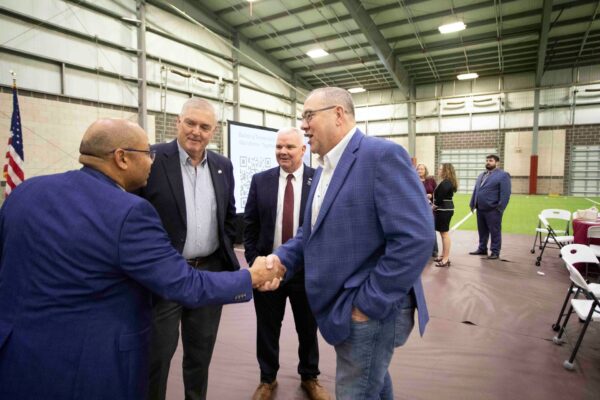 Four men in business attire stand inside a large indoor facility, with two of them in the foreground shaking hands and smiling. Others, including a woman, are visible in the background near chairs and tables.