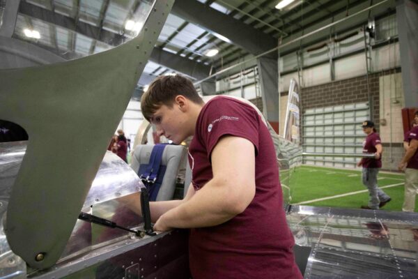 A young person in a maroon shirt works on assembling an aircraft inside a large indoor facility, while others in similar shirts are visible in the background.