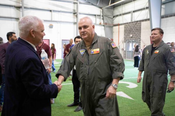 A man in a flight suit shakes hands with another man in a business suit inside an indoor facility with artificial turf, while other people stand and walk in the background.