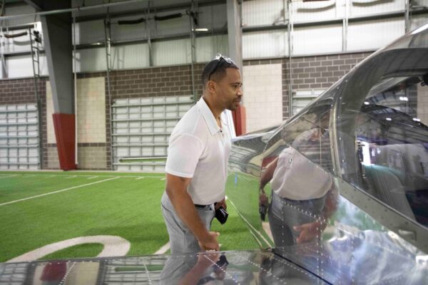 A man in a white polo shirt looks closely at the side of a silver airplane inside a large indoor facility with artificial turf and a brick wall.