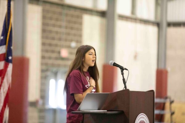 A young woman speaks passionately at a podium with a microphone indoors, standing next to an open laptop. An American flag is visible to the left in the background.
