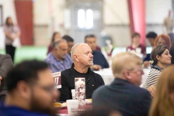 A group of people sit at round tables during an indoor event. The focus is on a bald man in a black shirt looking ahead, while others around him listen attentively. The background is softly blurred.