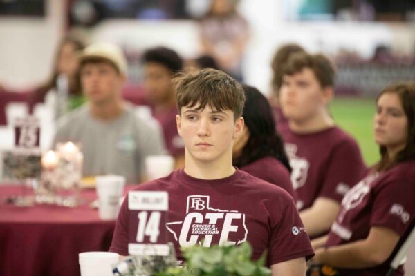 A young person wearing a maroon shirt with CTE and FB sits at a table among other students, all attentively listening at an indoor event. The atmosphere is focused and academic.