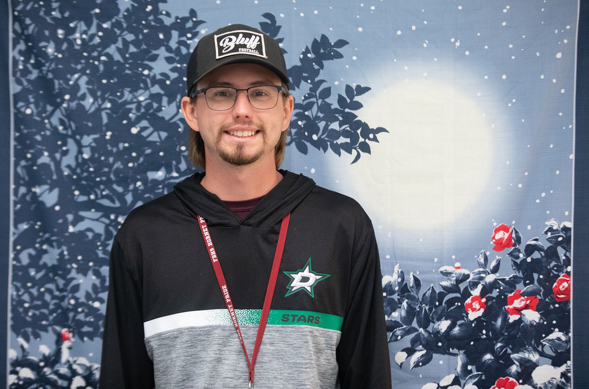 Josh Savins - Audio & Video Production A smiling man wearing glasses, a black hat, and a Dallas Stars hoodie stands in front of a backdrop with a full moon, tree branches, and red and white flowers.