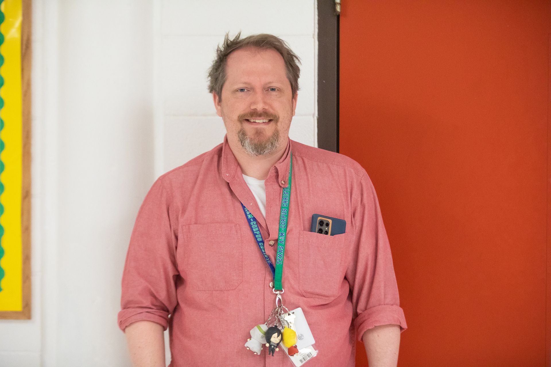 Joshua Blankenship - Arts, Audio/Video Technology, and Communications A man with a beard wearing a pink button-up shirt stands indoors, smiling. He has lanyards and keys around his neck and a smartphone in his shirt pocket. A red door and a white wall are in the background.
