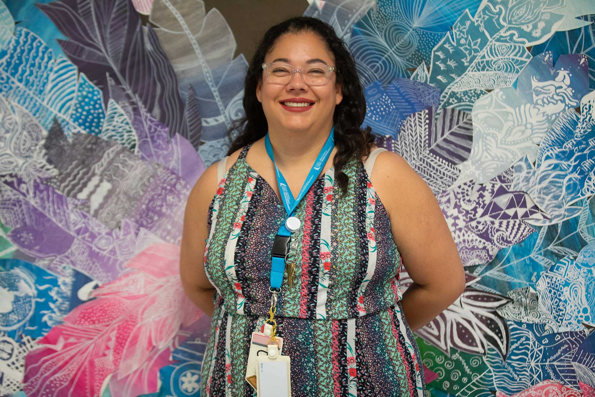 Julia-Tamez-Principles-of-IT- A woman wearing glasses and a colorful patterned dress stands smiling in front of a vibrant, abstract feathered background. She has a blue lanyard with various badges and keys around her neck.