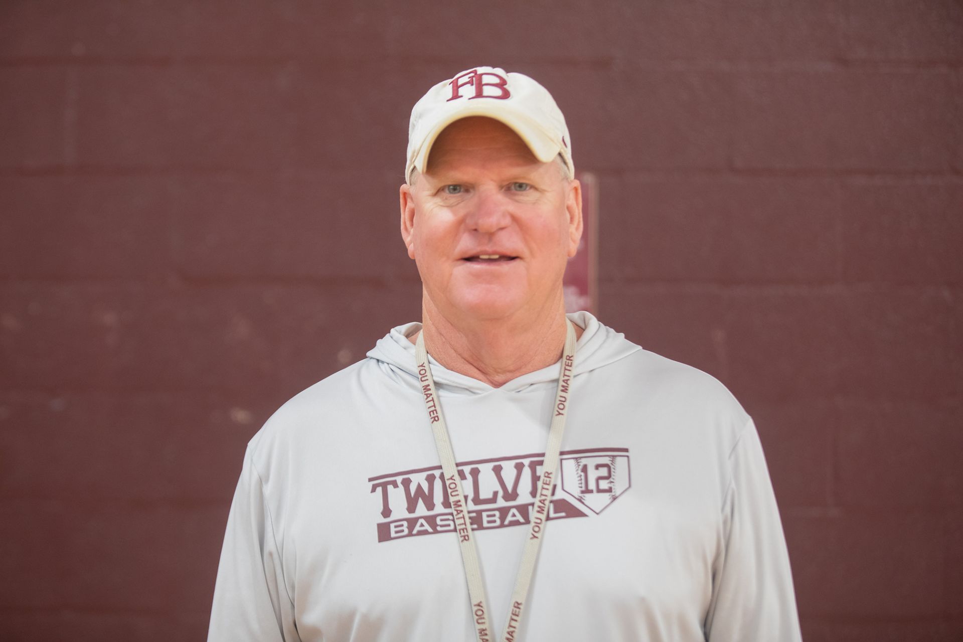 Kevin Nix - Principles of Construction A man wearing a white Twelve Baseball hoodie, a white cap with TB logo, and a lanyard stands in front of a maroon brick wall, looking at the camera and smiling slightly.