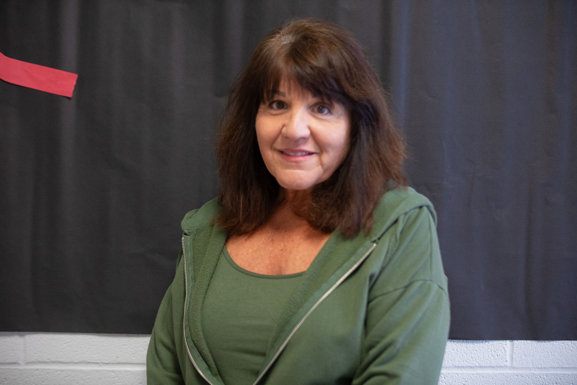 Linda Millard - Health Science A woman with medium-length brown hair wearing a green top and zip-up jacket is smiling at the camera. She is standing in front of a dark background with a red diagonal stripe.