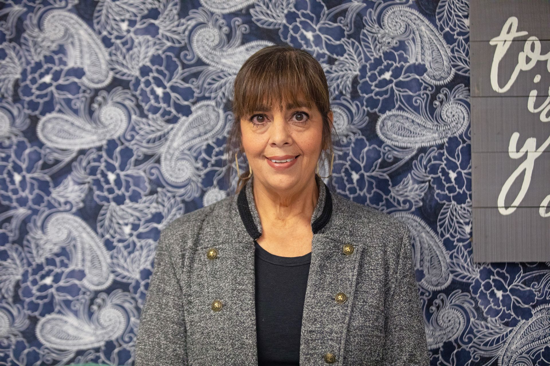 Liza Garza - BIM Financial / Math CTE Chair A woman with brown hair and bangs, wearing a gray jacket and black shirt, stands in front of a blue floral-patterned background and part of a gray sign with white cursive text.