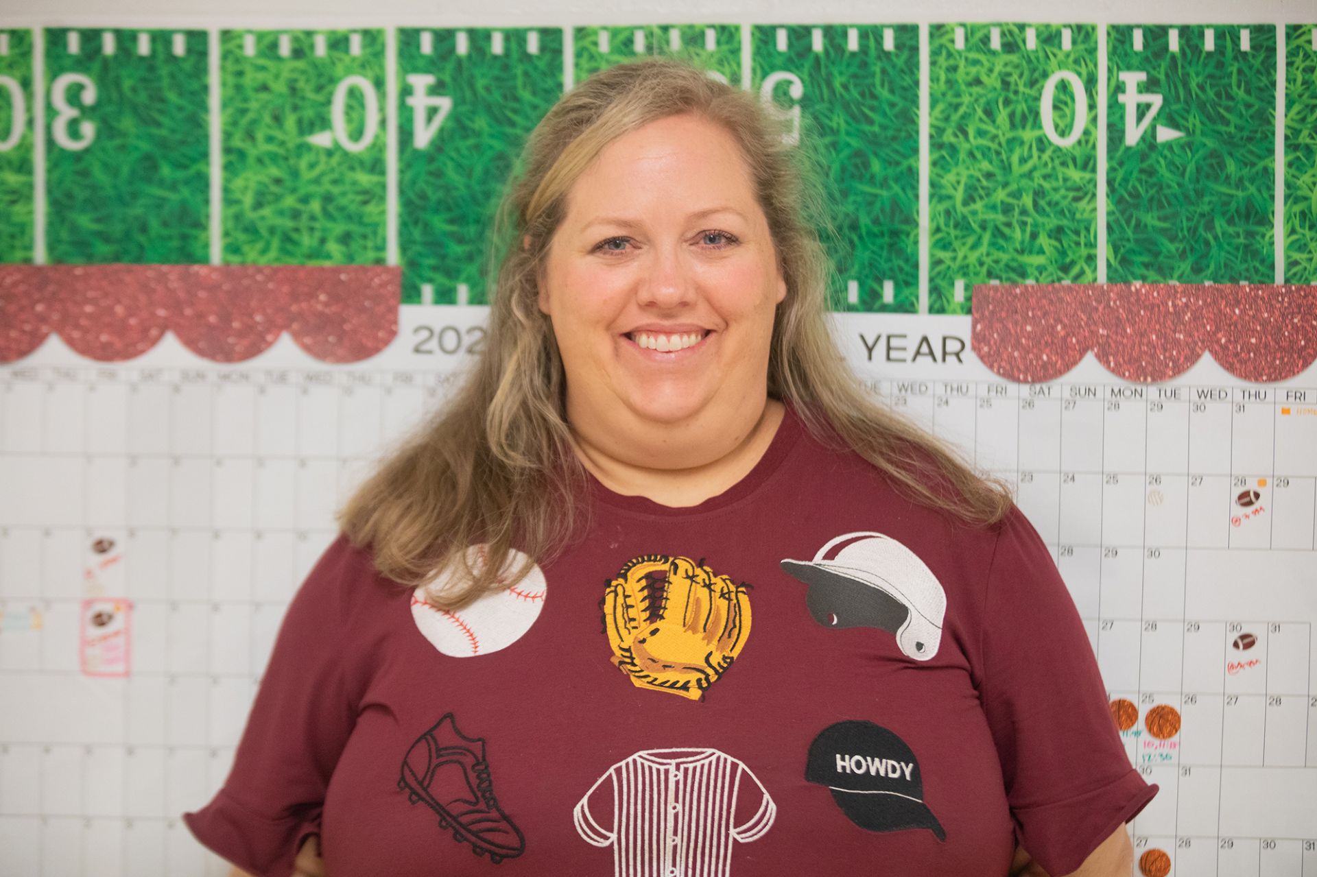 Rachel Johnson - Practicum of Entrepreneurship, Fashion Marketing, Advertising A woman with long blonde hair smiles in front of a calendar and a football-themed bulletin board. She is wearing a maroon shirt with sports-themed graphics, including a baseball, glove, helmet, cleat, jersey, and a patch that says HOWDY.