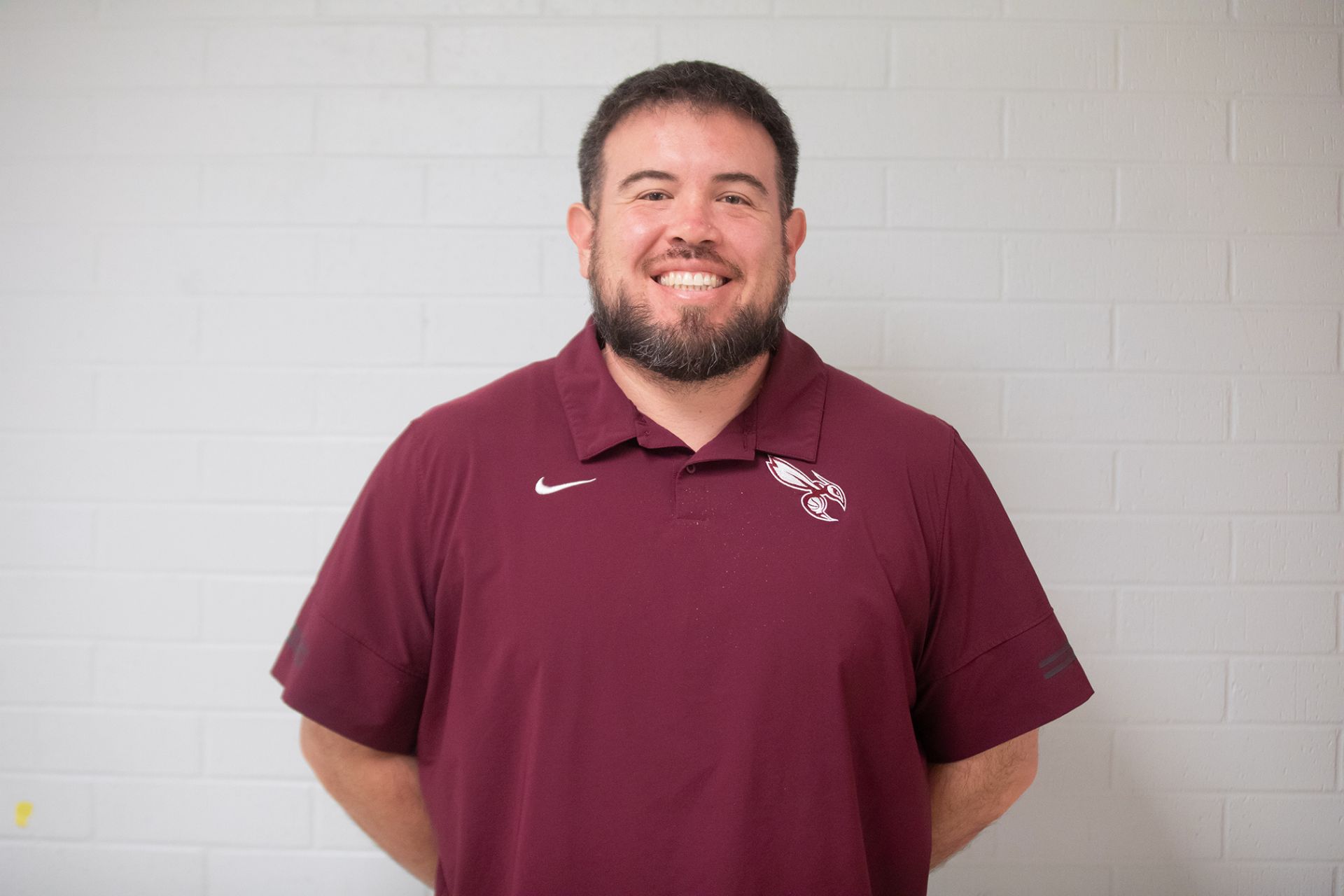 Richard Cervantes - Principles of Transportation A man with short dark hair and a beard smiles at the camera, wearing a maroon collared shirt with a white logo. He stands in front of a light gray brick wall with his hands behind his back.