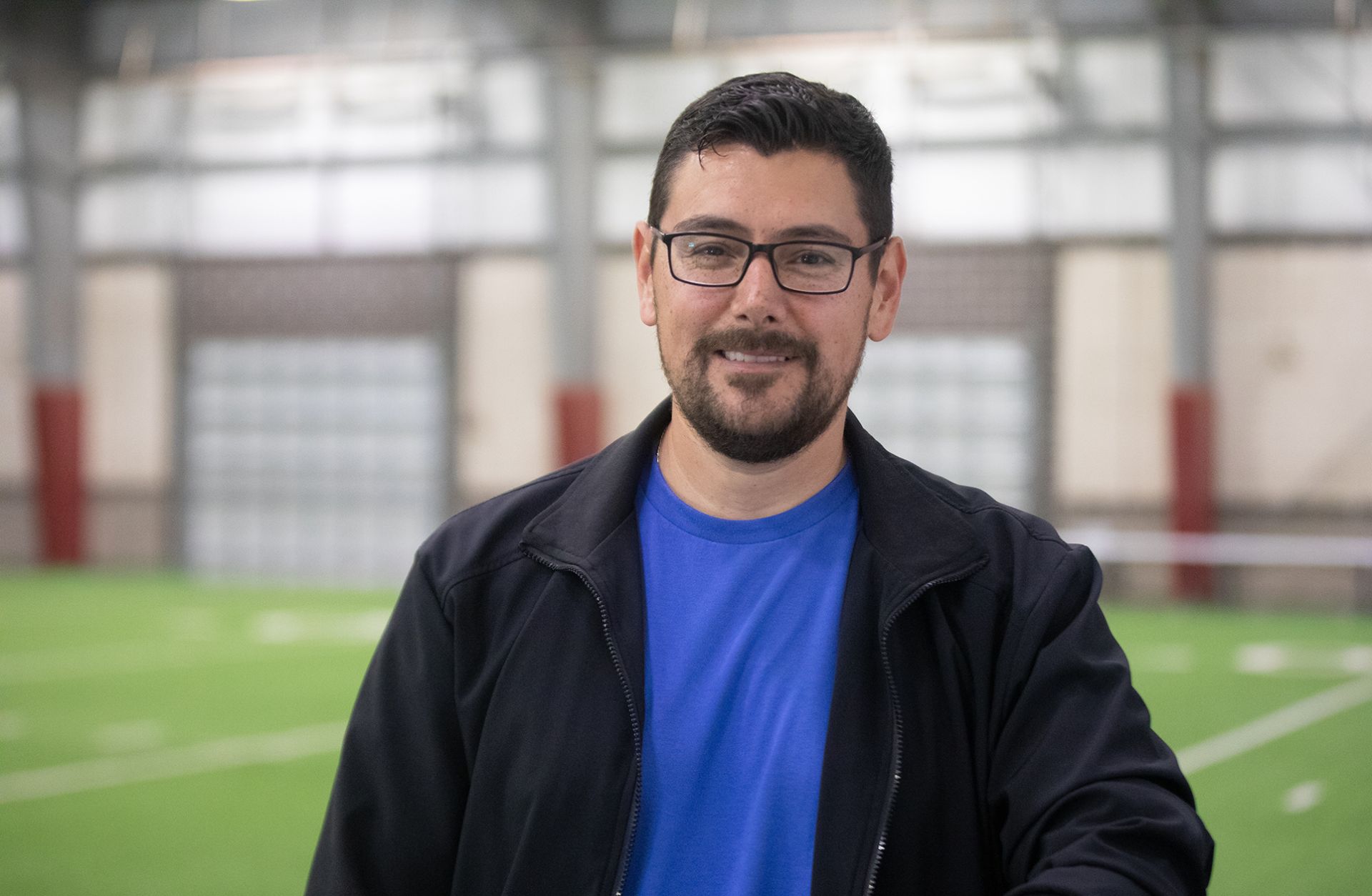 Roland Medrano - Hornet Aviation / Drone A man with short dark hair, glasses, and a beard is smiling while standing indoors on artificial turf. He is wearing a blue shirt and a black jacket, with a blurred sports facility in the background.