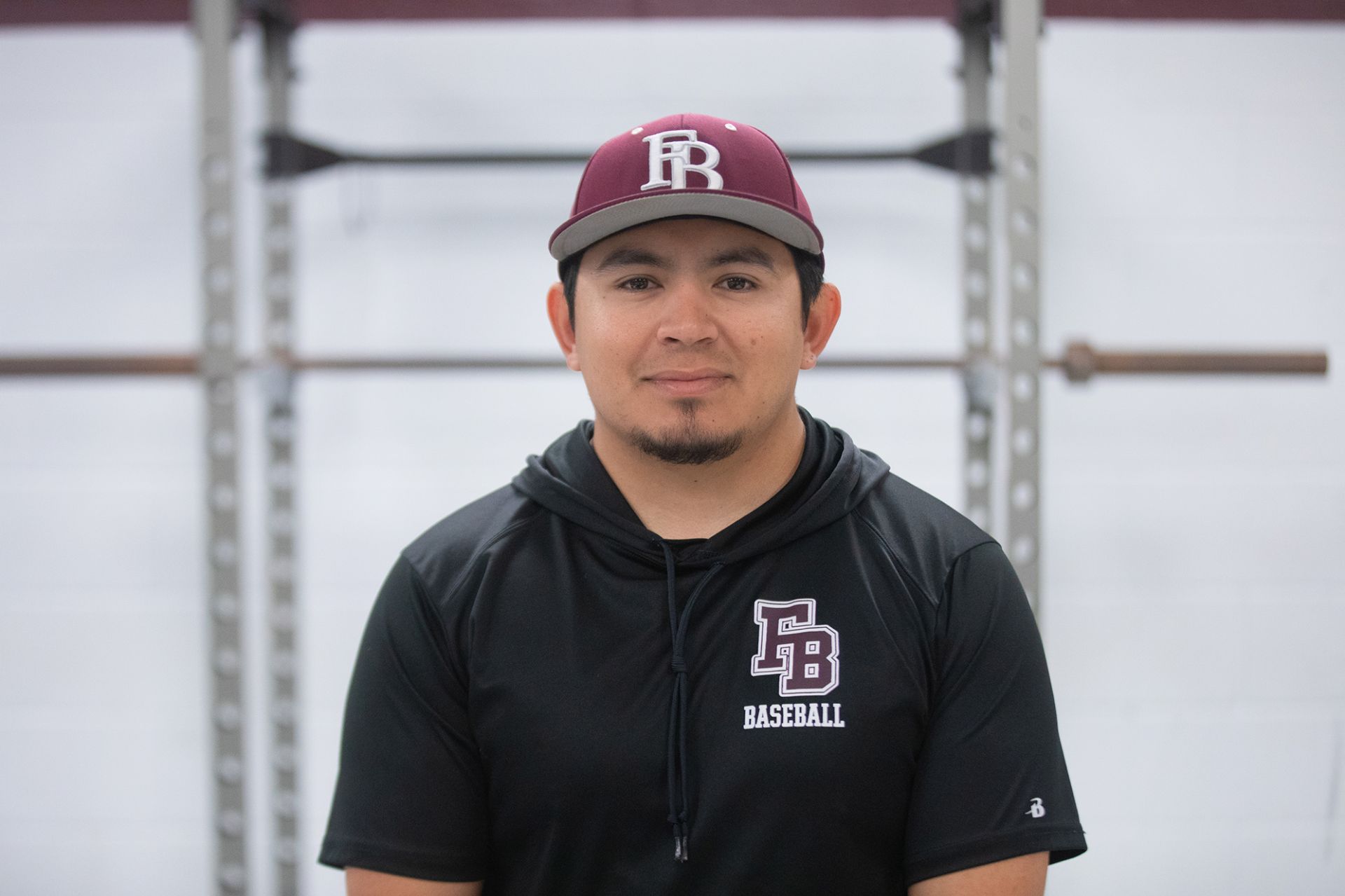 Sam Quintana - Construction A man wearing a maroon baseball cap and a black hoodie with EB Baseball on it stands in front of gym equipment, looking at the camera with a neutral expression.