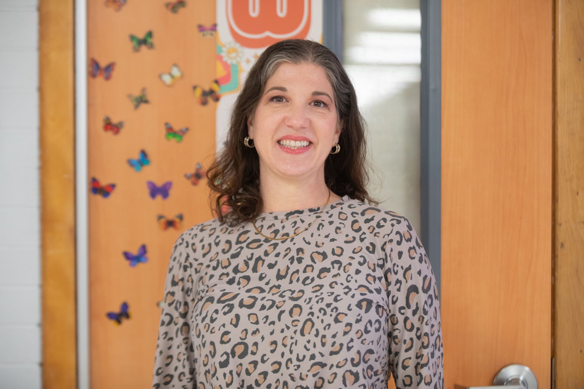Sarah Laza - Professional Communications A woman with wavy brown hair wearing a leopard print blouse stands smiling in front of a classroom door decorated with colorful butterfly cutouts.