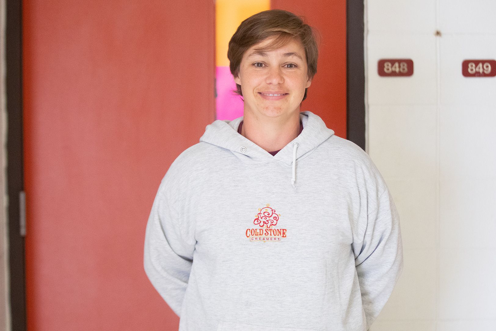 Shannon Dana - Health Science, College & Career Exploration A person with short hair wearing a light gray Cold Stone Creamery hoodie stands indoors, smiling in front of a red door with room numbers 848 and 849 visible on the white wall.