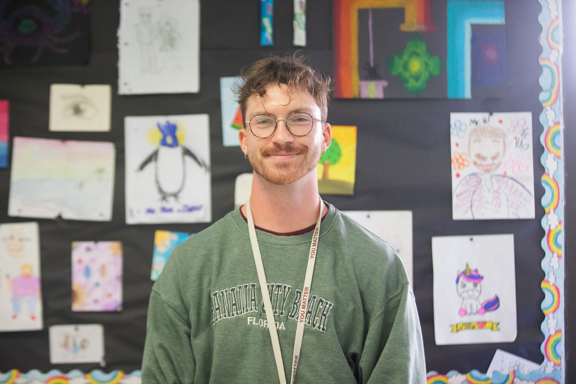 Tanner Killingsworth - Business Marketing & Finance A young man with glasses and a beard smiles in a classroom, wearing a green Daytona Beach, Florida sweatshirt and a lanyard. Behind him, colorful childrens drawings are displayed on a bulletin board.