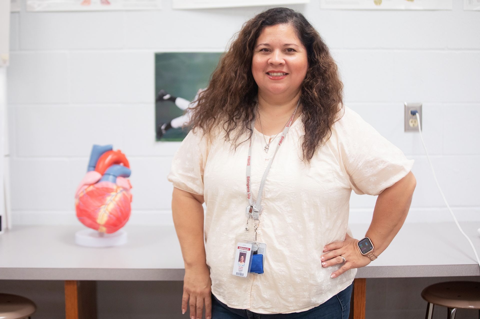 Valerie Garcia - Health Science A woman with curly hair wearing a white shirt and an ID badge stands in a classroom. Behind her, there is a model of a human heart on a counter and science posters on a white wall.