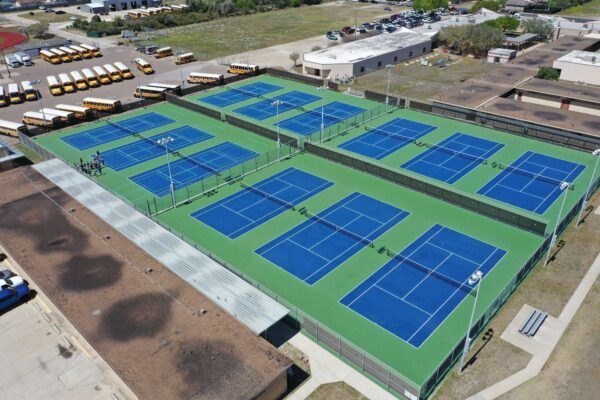 Aerial view of a sports complex with twelve blue and green tennis courts, surrounded by fencing. School buses are parked nearby, and groups of people can be seen on one side of the courts. Buildings and open fields are in the background.