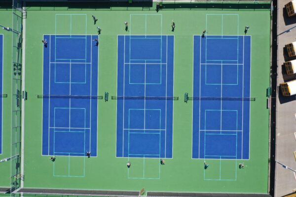 Aerial view of three blue tennis courts with green borders. Several players are on each court, and yellow school buses are parked near the courts on the right side of the image.