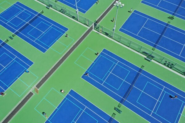 Aerial view of several people playing tennis on large, blue and green outdoor courts, separated by fences and pathways, under bright daylight.