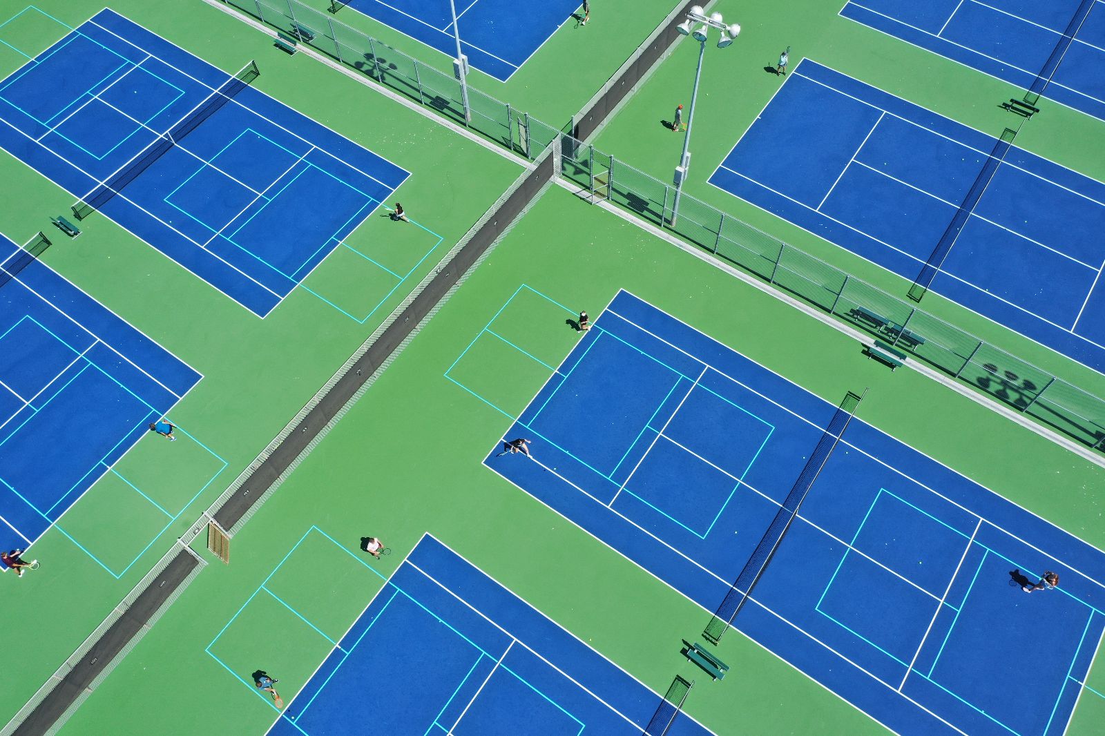 Aerial view of several people playing tennis on large, blue and green outdoor courts, separated by fences and pathways, under bright daylight.