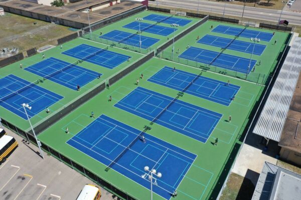 Aerial view of an outdoor tennis facility with eight blue and green courts, several people playing on each court, and surrounding fencing. Nearby are buildings, a road, and parked school buses.