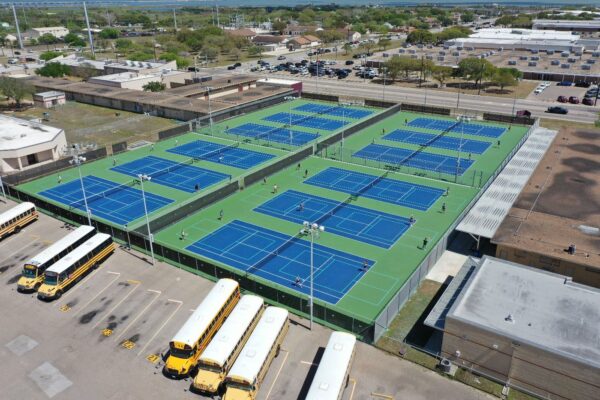 Aerial view of a school tennis complex with multiple blue courts, several people playing, and yellow school buses parked nearby. Surrounding buildings and parking lots are visible in the background.