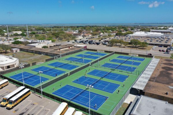 Aerial view of a large outdoor tennis facility with multiple blue and green courts, surrounded by school buses, buildings, and parking lots under a clear blue sky. Players are visible on several courts.
