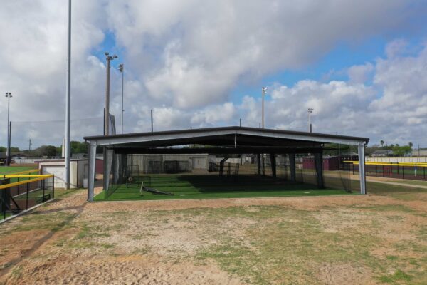 A covered outdoor baseball or softball batting cage with netting, artificial turf, and pitching machines, located in an athletic field under a cloudy sky.