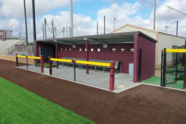 A maroon and gray baseball dugout with benches and metal roof, surrounded by a chain-link fence with yellow padding, next to a brown dirt field and green artificial turf, under a partly cloudy sky.