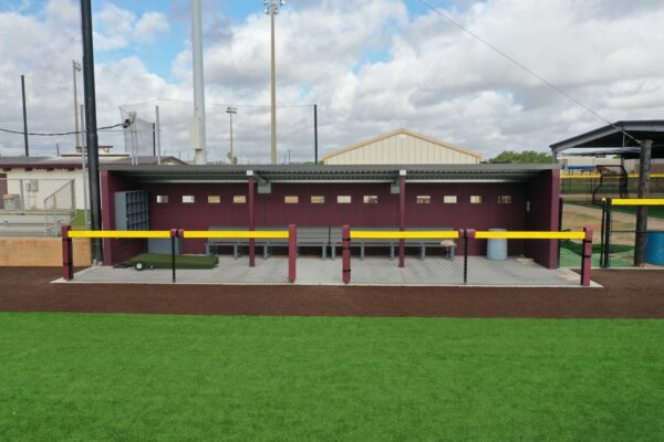 A baseball dugout with maroon walls, a metal roof, benches, and storage areas is behind a chain-link fence with yellow padding. The area is surrounded by artificial turf and nearby sports fields.