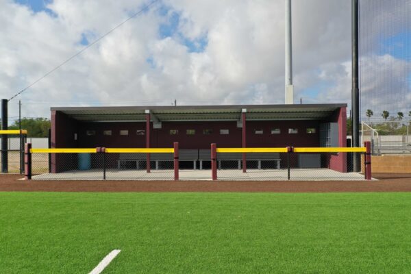 A maroon baseball dugout with a gray roof, benches inside, and a yellow rail in front, situated behind a chain-link fence on a field with bright green artificial turf.