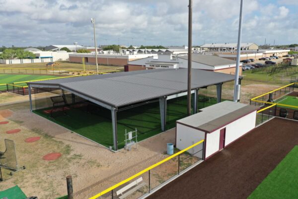 A covered outdoor batting cage with artificial turf, adjacent to a brown and white shed, sits next to a fenced baseball field. Nearby buildings and parked cars are visible in the background under a partly cloudy sky.