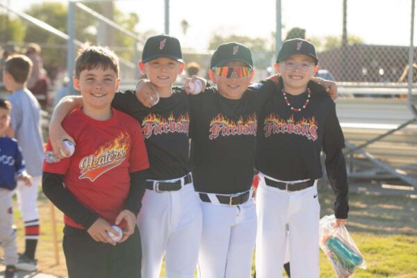 Four young boys pose on a sunny baseball field. Three wear matching Fireballs uniforms and baseball caps, with arms around each other, and one in a red Heaters shirt stands beside them, all smiling at the camera.