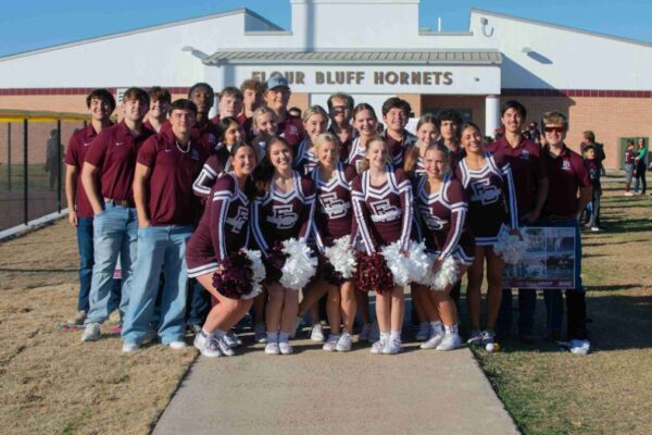 A group of high school cheerleaders and students in maroon uniforms pose and smile together outside a building labeled Bluff Bluff Hornets on a sunny day.