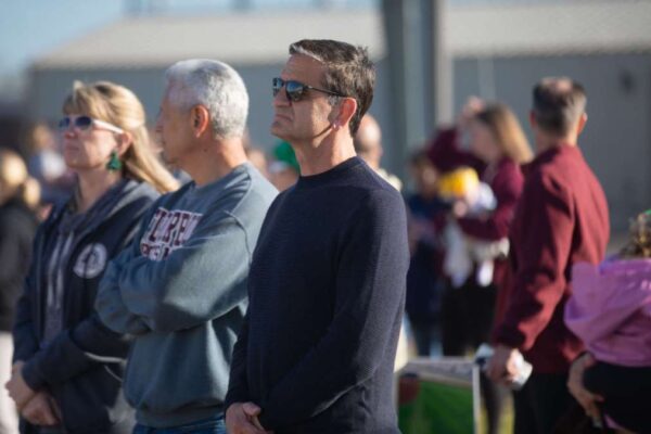 A man in sunglasses stands outdoors among a group of people, looking forward with a serious expression. Other individuals stand nearby, some blurred in the background, all appearing to watch something off-camera.