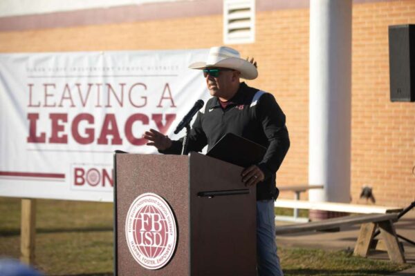 A man in a cowboy hat speaks at a podium outdoors, with a microphone and folder. Behind him, a banner reads Leaving a Legacy. The event appears to be at a school or district building.