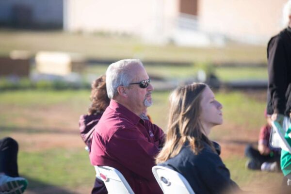 A man wearing sunglasses and a maroon shirt sits outdoors on a white chair, alongside a young woman, both looking forward attentively. The background is blurred, suggesting an outdoor event.