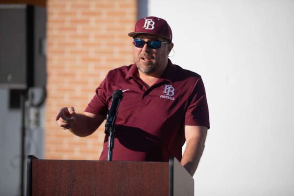 A man wearing sunglasses, a maroon baseball cap, and a matching LB Football shirt speaks into a microphone at an outdoor podium, gesturing with one hand. A brick wall and white background are visible behind him.
