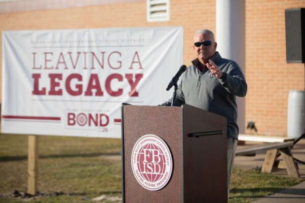 A man in sunglasses speaks at a podium outdoors in front of a large sign that reads Leaving a Legacy and BOND. The podium has a logo for a school district. The background includes a brick building.