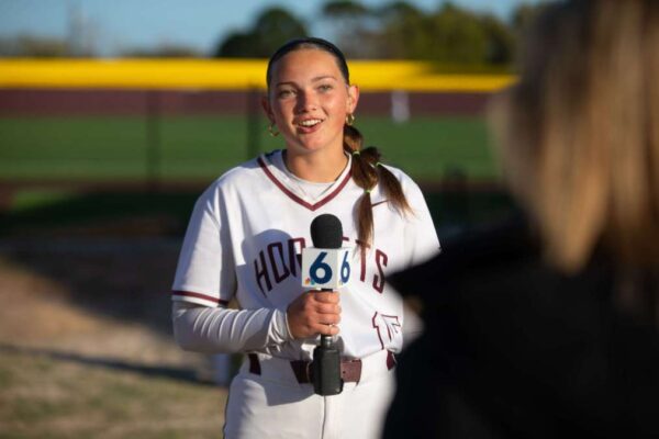 A softball player in uniform is being interviewed on a field, holding a microphone labeled with the number 6, while a reporter stands in the foreground with their back to the camera.