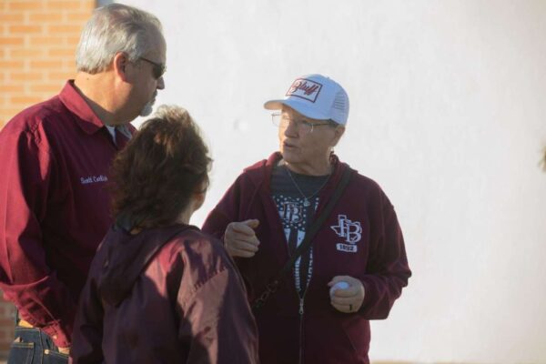 Three people are talking outdoors. Two men and one woman wear maroon jackets; one man has a white beard and sunglasses, and the other wears glasses and a white baseball cap. The group stands near a white wall.