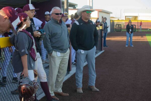 Two men stand by a baseball field dugout observing a game, one in a light suit and sunglasses, the other in a cowboy hat and boots. Several players in maroon uniforms and hats stand nearby, some leaning on the fence.