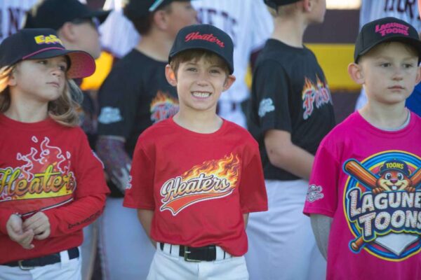Three young children in colorful baseball uniforms stand in a row, with a smiling boy wearing a red Heaters shirt in the center, flanked by a girl in a yellow Heaters shirt and a boy in a pink Laguna Toons shirt.