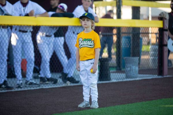 A young baseball player in a yellow shirt and white pants stands on the field holding a ball, with a team in white uniforms watching from behind a fence in the background.
