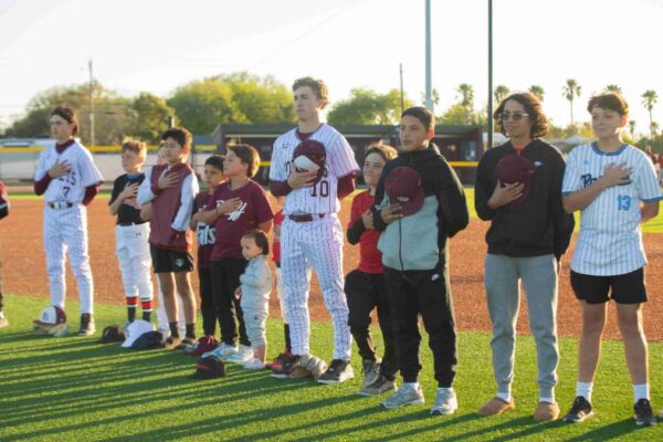 A group of young baseball players and children stand in a line on the field with their right hands over their hearts, some holding baseball caps, during a pre-game ceremony at sunset.